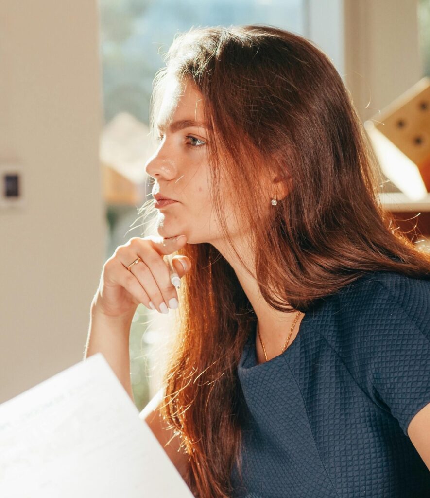 A thoughtful woman looks out the window while holding papers, bathed in warm sunlight indoors.